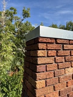 Close-up of a brick chimney corner with a new galvanized cap edge and oak foliage behind it, Osprey FL